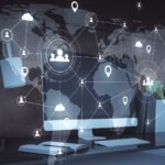 A monitor on a desk against a world-map wall, overlaid with a glowing network of connected nodes, people, and location pins across the continents
