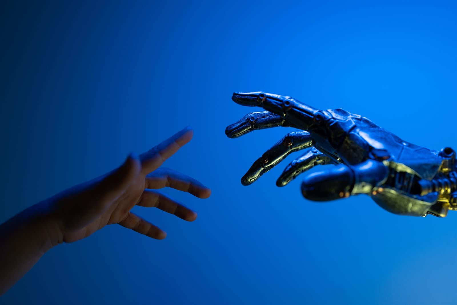 A human hand and a metallic robotic hand reaching toward each other against a deep blue background, fingertips almost touching