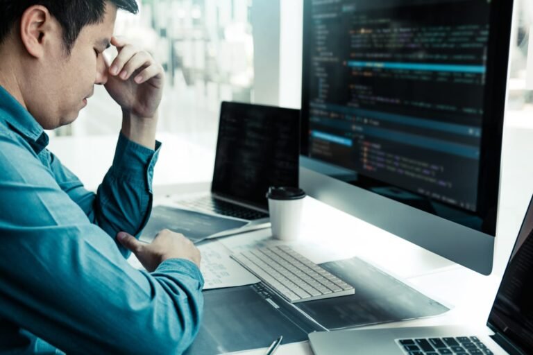 A software developer sits back from his desk with his eyes closed and his hand pressed against the bridge of his nose, a monitor full of code in front of him.